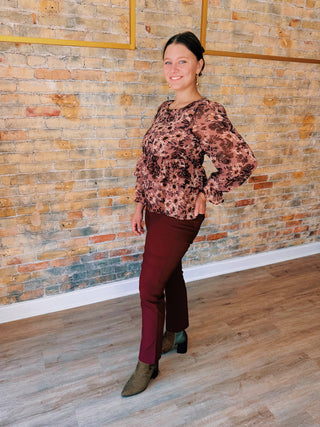 Woman in a floral blouse and burgundy pants standing against a brick wall.