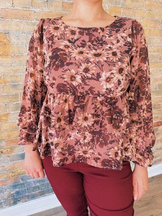 Person wearing a floral blouse with long sleeves and maroon pants against a brick wall.