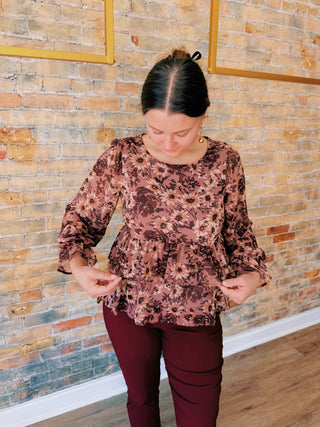 Woman wearing a floral blouse and maroon pants against a brick wall.