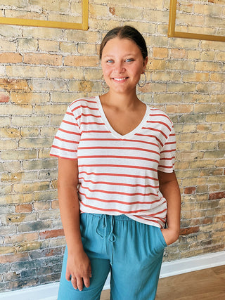 Person wearing a striped white & red t-shirt  and blue pants standing against a brick wall.