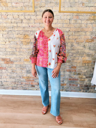 Woman wearing a colorful patterned blouse and blue jeans standing against a brick wall.