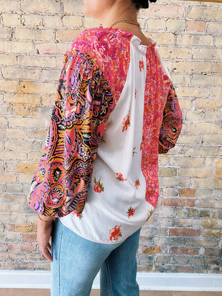 Person wearing a colorful patterned top with a white floral shirt underneath against a brick wall.