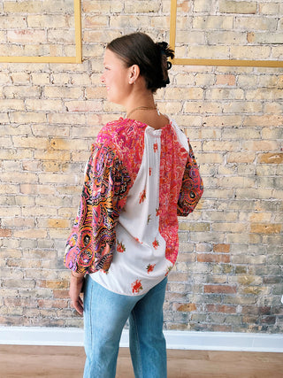 Woman wearing a colorful patterned top and carrying a white tote bag with floral designs against a brick wall.