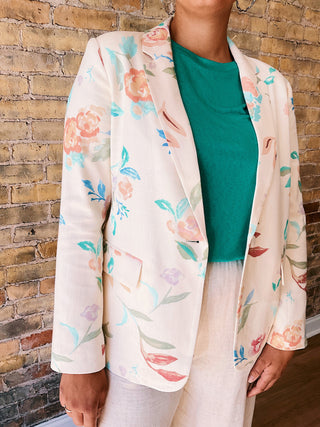 Person wearing a floral kimono-style jacket over a green top and white pants against a brick wall.