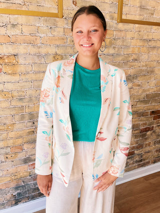 Woman wearing a light-colored floral blazer over a green top and white pants, standing against a brick wall.