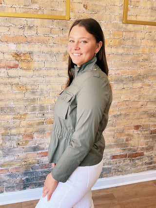 Woman wearing a green jacket and white pants standing against a brick wall.