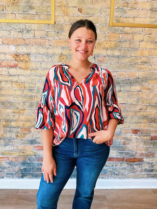 Woman wearing a colorful patterned blouse and blue jeans standing against a brick wall.