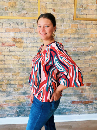 Woman wearing a colorful patterned blouse and blue jeans standing against a brick wall.