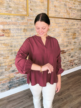 Woman wearing a burgundy blouse and white pants standing against a stone wall.