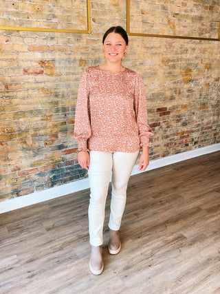 A woman standing against a brick wall backdrop wearing a pink animal print blouse with long sleeves and beige pants.