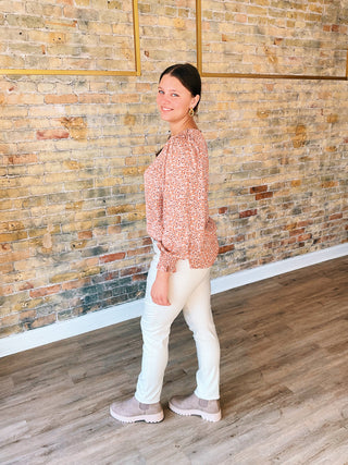 Woman standing against a brick wall wearing a patterned top and white pants.