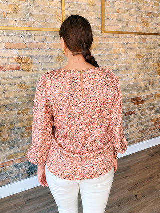 Woman wearing a pink floral blouse and white pants against a stone wall.