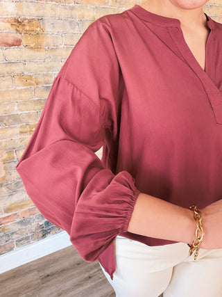 A person wearing a dark ginger colored top with a round neckline and 3/4 sleeves, paired with light-colored pants, standing in front of a brick wall.