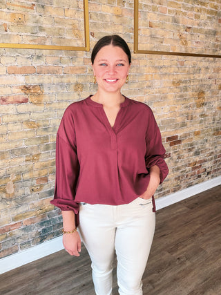 Woman wearing a maroon blouse and white pants standing against a brick wall.