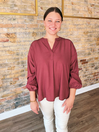 Woman wearing a burgundy blouse and white pants standing against a brick wall.