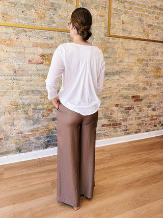 Woman wearing a white blouse and brown pants standing against a brick wall.