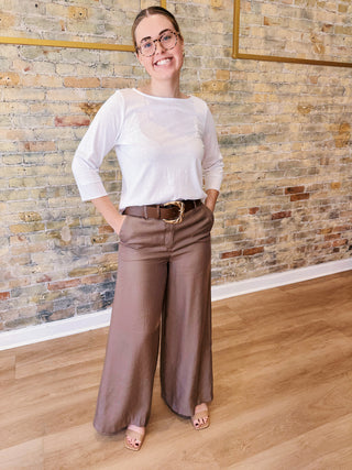 Woman wearing a white top and brown pants standing in front of a brick wall.