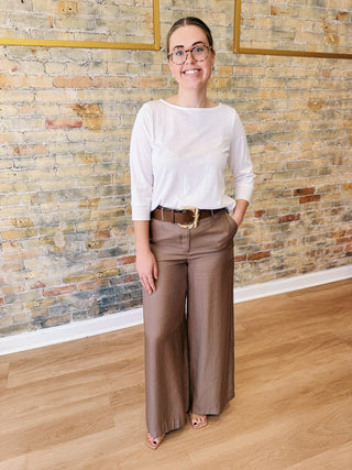 Woman wearing a white sweater and brown pants standing in front of a brick wall.