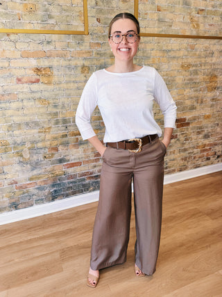 Woman wearing a white shirt and brown pants standing in front of a brick wall.