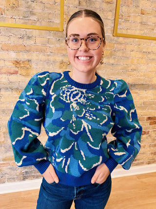 Person wearing a blue floral patterned sweater in front of a wooden paneled wall.