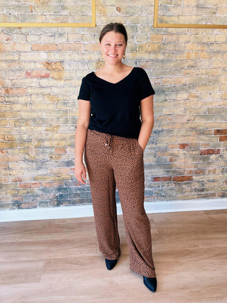 Model wearing Callie Pants — wide-leg brown animal print trousers with elastic waist, paired with black top and black boots, standing against brick wall background.