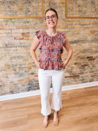 Woman wearing a floral blouse and white pants standing against a brick wall.