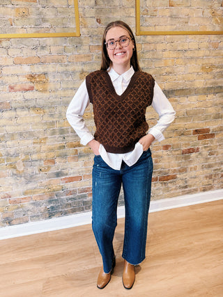 Person wearing a brown knitted vest over a white shirt and blue jeans, standing against a stone wall.