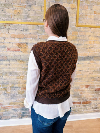 Person wearing a brown patterned vest over a white shirt and blue jeans against a brick wall.