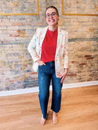 Person wearing a floral blazer, red shirt, and blue jeans standing against a brick wall.
