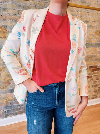 Person wearing a floral blazer over a red shirt and blue jeans against a brick wall.