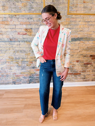Woman wearing a floral blazer, red shirt, and blue jeans standing against a brick wall.