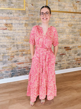 Woman in a pink floral dress standing against a brick wall.
