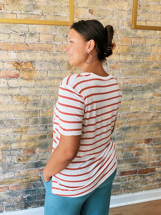 Person wearing a striped white & red t-shirt  and blue pants standing against a brick wall.
