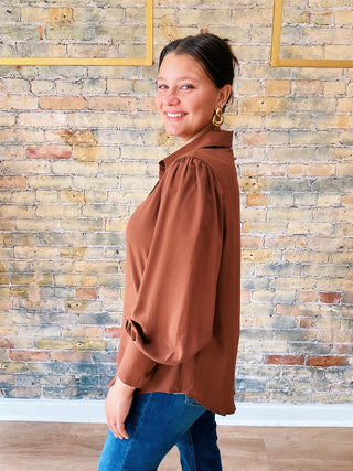 Woman wearing a brown blouse and blue jeans standing against a brick wall.