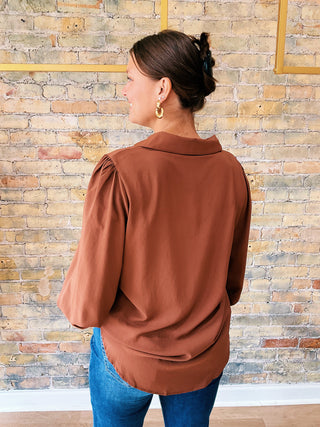 Woman wearing a brown blouse and blue jeans against a brick wall.