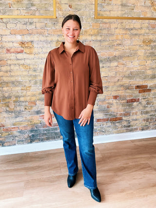 Woman wearing a brown shirt and blue jeans standing against a brick wall.