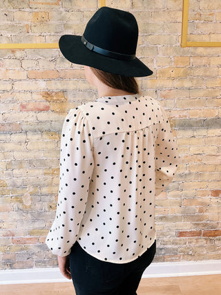 Person wearing a black hat and polka dot blouse against a brick wall.