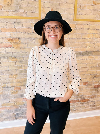 Person wearing a polka dot blouse and black hat against a brick wall.