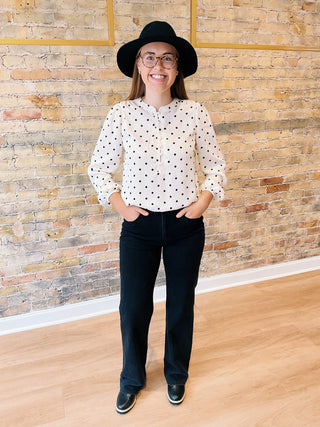 Person wearing a polka dot blouse, black pants, and a hat against a brick wall.