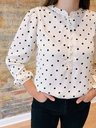 White blouse with black polka dots worn by a person against a brick wall.