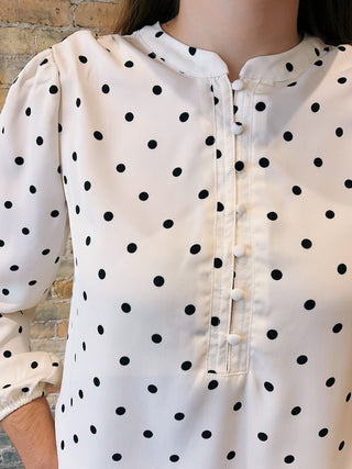 White blouse with black polka dots worn by a person against a brick wall.