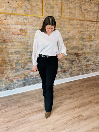 Woman in a white blouse and black pants standing against a brick wall.
