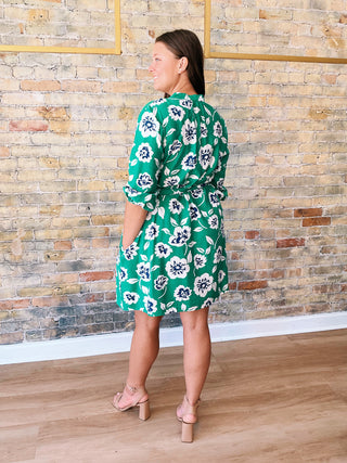 Woman wearing a green floral dress standing against a brick wall.