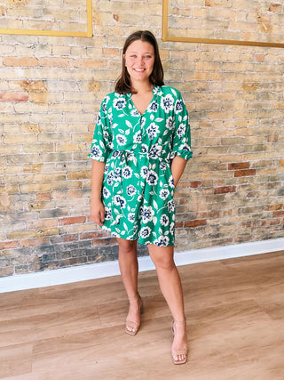 Woman wearing a green floral dress standing against a brick wall.