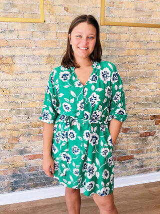 Woman wearing a green floral dress standing against a brick wall.