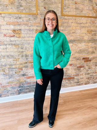 Person wearing a green sweater and black pants standing against a brick wall.