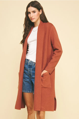 Woman wearing a long rust-colored coat over a white shirt and denim shorts on a beige background