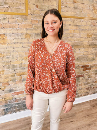 A woman wearing a long sleeve orange and white floral crossover top with beige pants, standing against a brick wall backdrop.