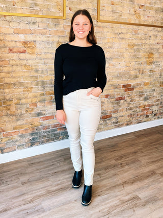 A person standing against a brick wall backdrop, wearing Faith brand straight pants with a textured, subtle metallic shine, in a beige color.