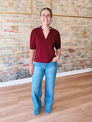 Person wearing a maroon shirt and blue jeans standing against a brick wall.